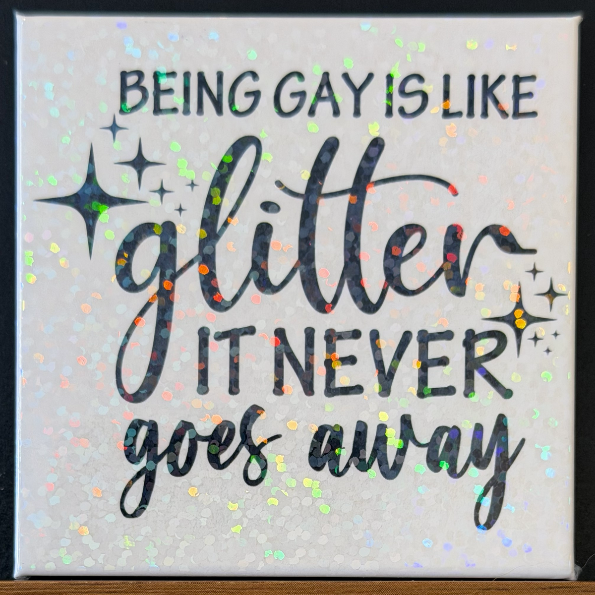 Decorative tile with text 'Being gay is like glitter, it never goes away' on a glittery white background.