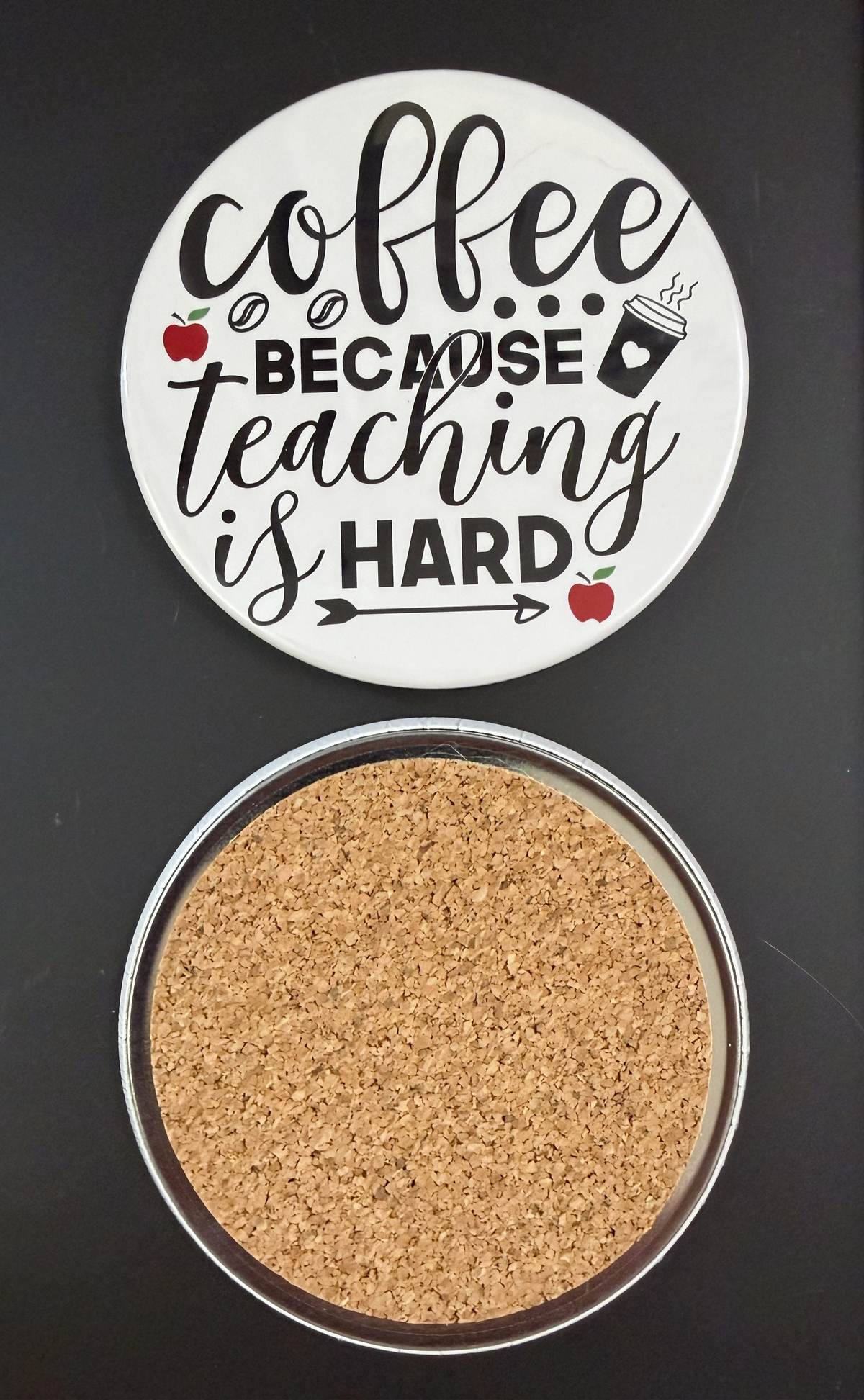 Round white drink coaster with cork backing with 'Coffee because teaching is hard' in black text, with small images of red apples, coffee beans and a coffee cup, photographed on a black background.
