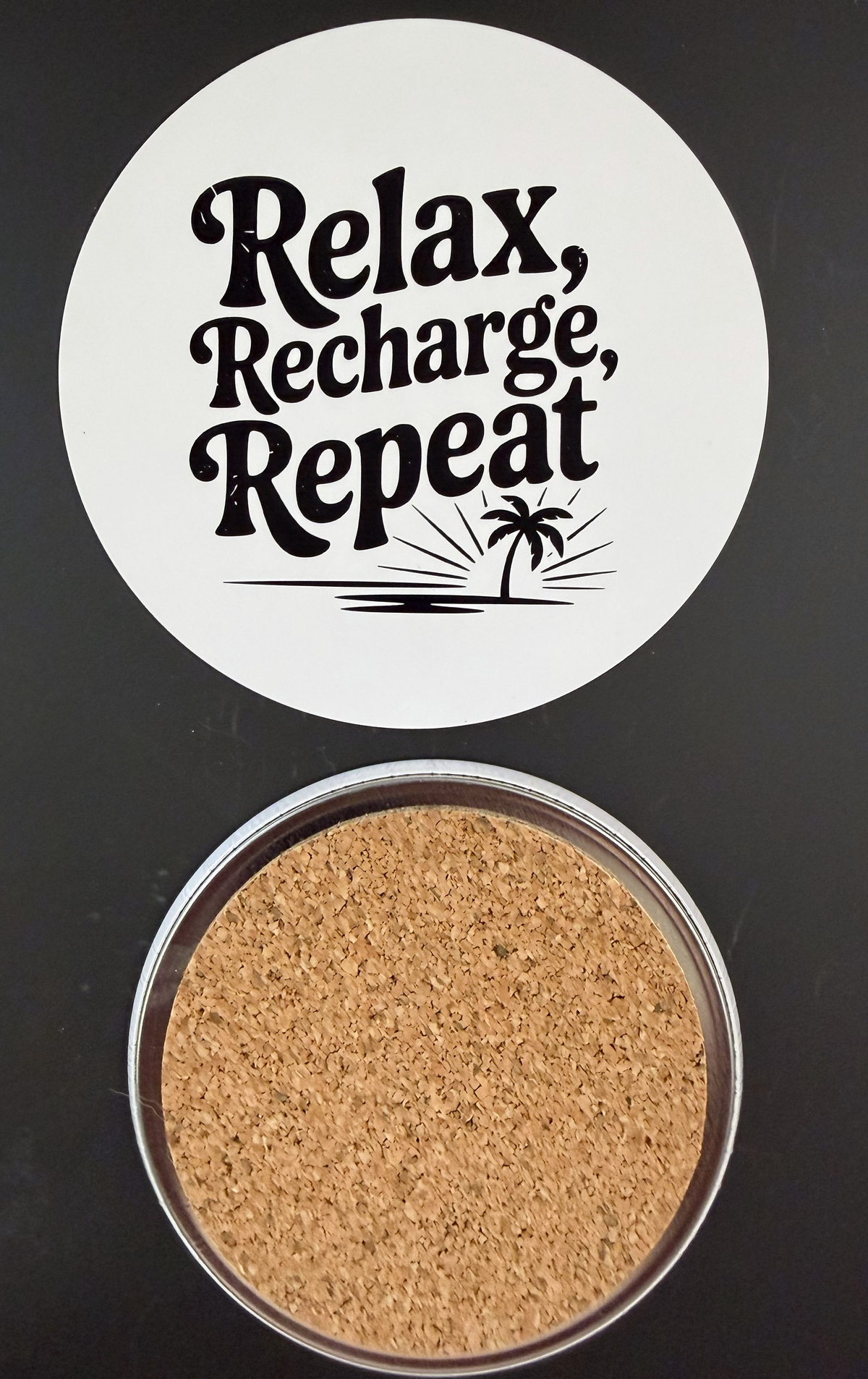 White round drink coaster with a cork backing with a black image of a palm tree and text 'relax, recharge, repeat' on a white background.  This photo was taken on a black background. 
