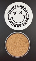 White round drink coaster with a cork backing with a black image of a smiley face with airplanes for eyes with text 'travel more, worry less' on a white background.  This photo was taken on a black background.