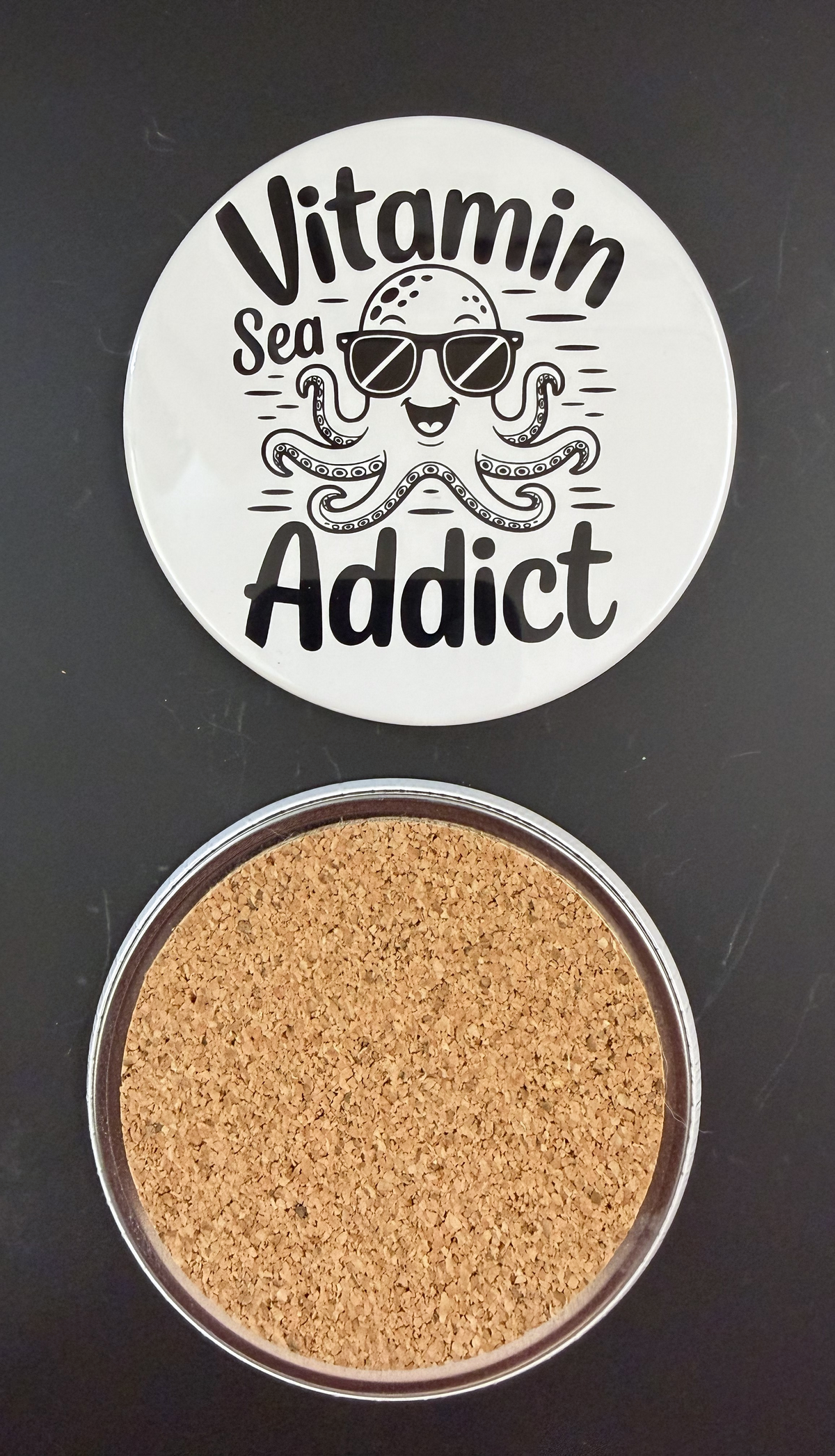White round drink coaster with a cork backing with a black image of a smiling octopus wearing sunglasses with text 'vitamin sea addict' on a white background.  This photo was taken on a black background.