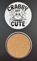 White round drink coaster with a cork backing with a black image of a smiling crab and seashells with text 'crabby but cute' on a white background.  This photo was taken on a black background.