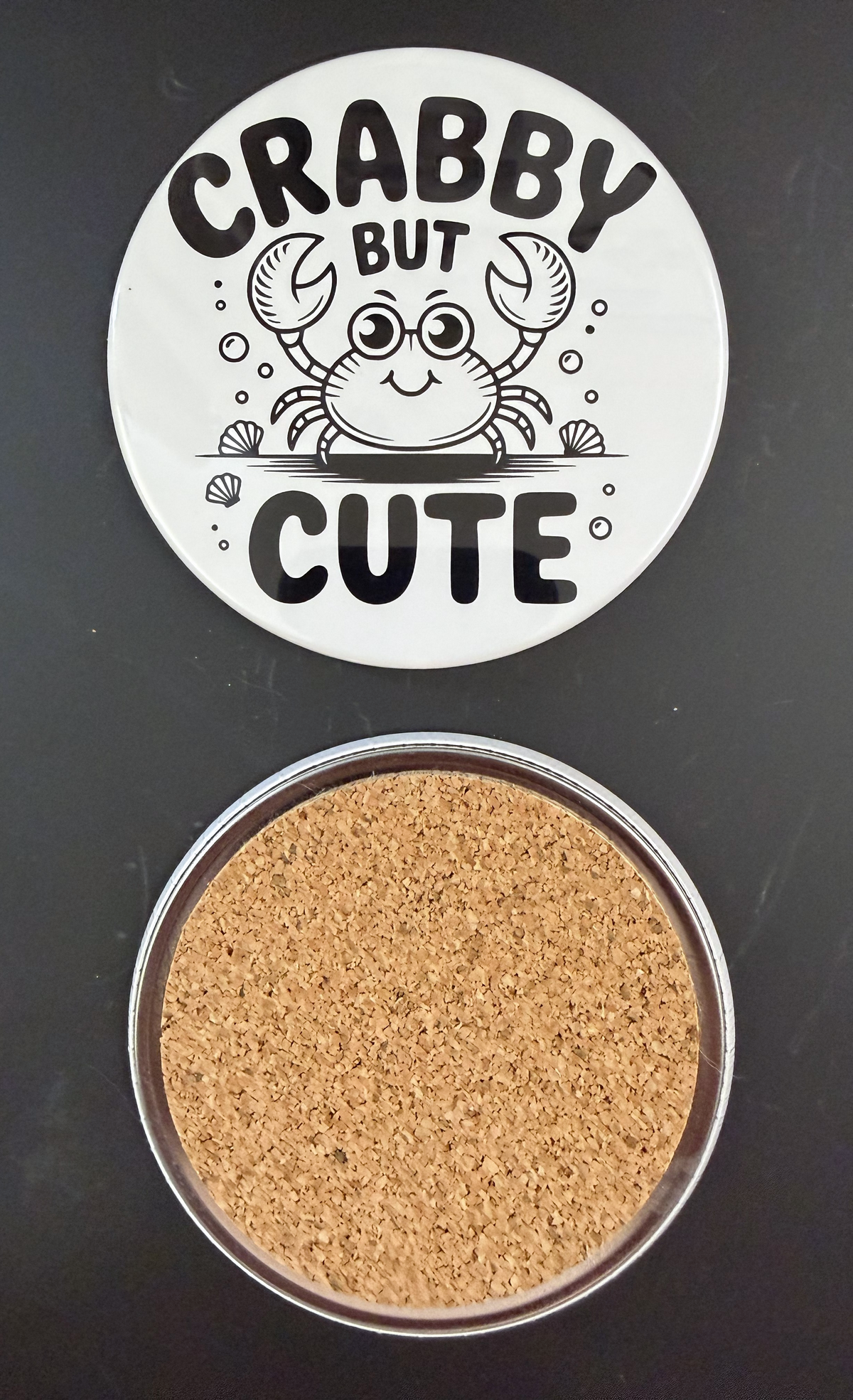 White round drink coaster with a cork backing with a black image of a smiling crab and seashells with text 'crabby but cute' on a white background.  This photo was taken on a black background.