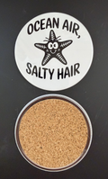 White round drink coaster with a cork backing with a black image of a smiling starfish with text 'ocean air, salty hair' on a white background.  This photo was taken on a black background.