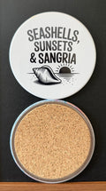 White round drink coaster with a cork backing with a black image of a sunset with a conch seashell on the beach with text 'seashells, sunsets and sangria' on a white background.  This photo was taken on a black background.
