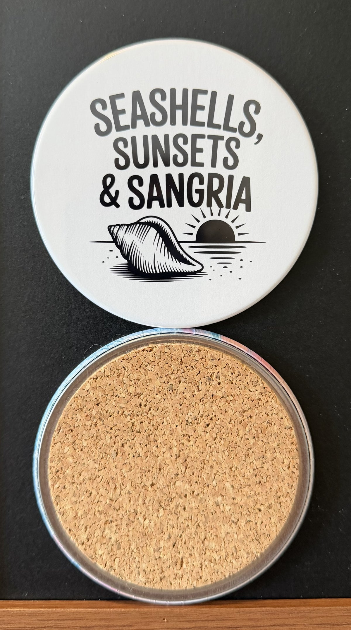 White round drink coaster with a cork backing with a black image of a sunset with a conch seashell on the beach with text 'seashells, sunsets and sangria' on a white background.  This photo was taken on a black background.