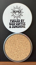 White round drink coaster with a cork backing with a black image of a smiling sun wearing sunglasses with text 'fueled by iced coffee & sarcasm' on a white background.  This photo was taken on a black background.