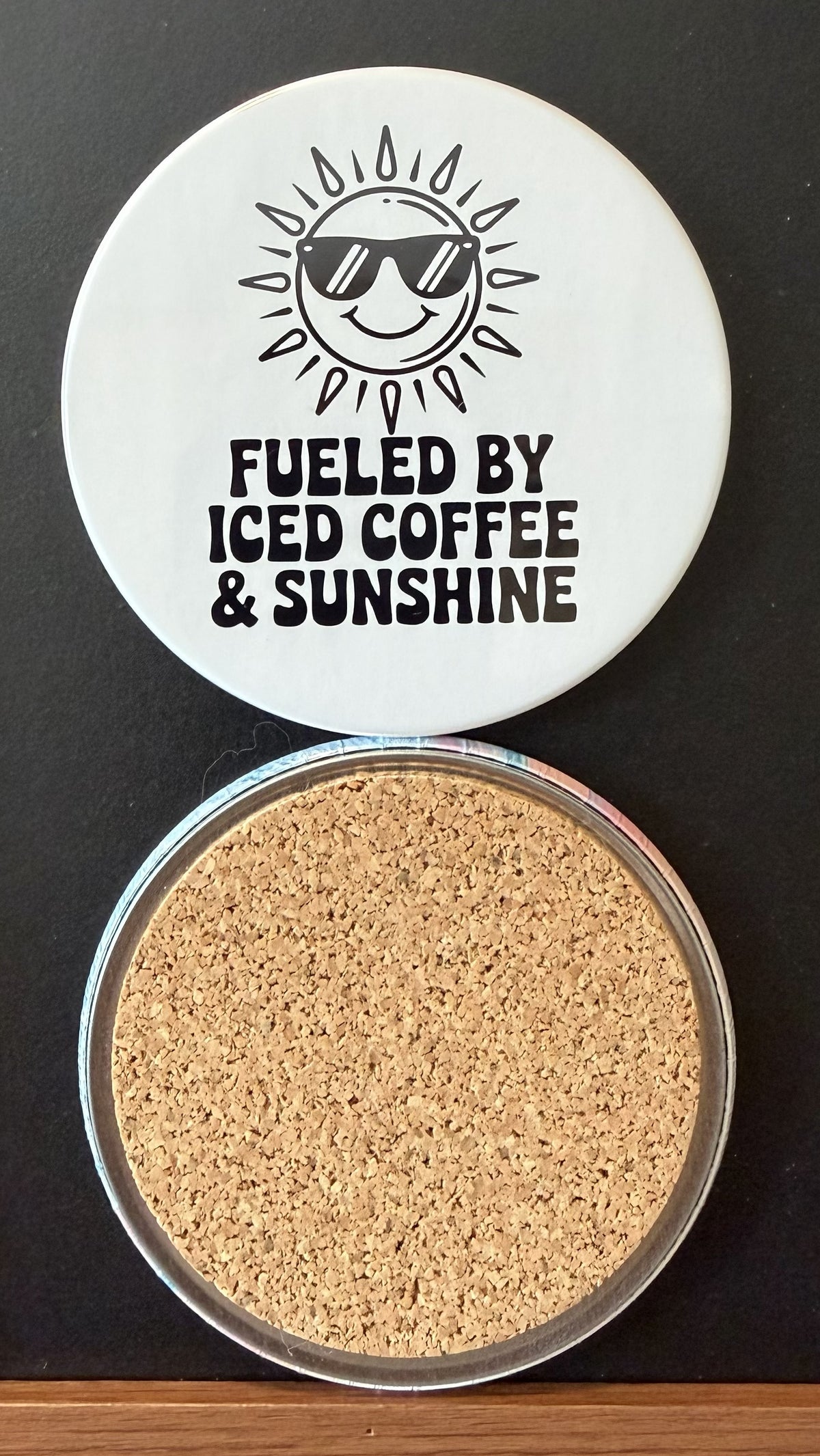 White round drink coaster with a cork backing with a black image of a smiling sun wearing sunglasses with text 'fueled by iced coffee & sarcasm' on a white background.  This photo was taken on a black background.