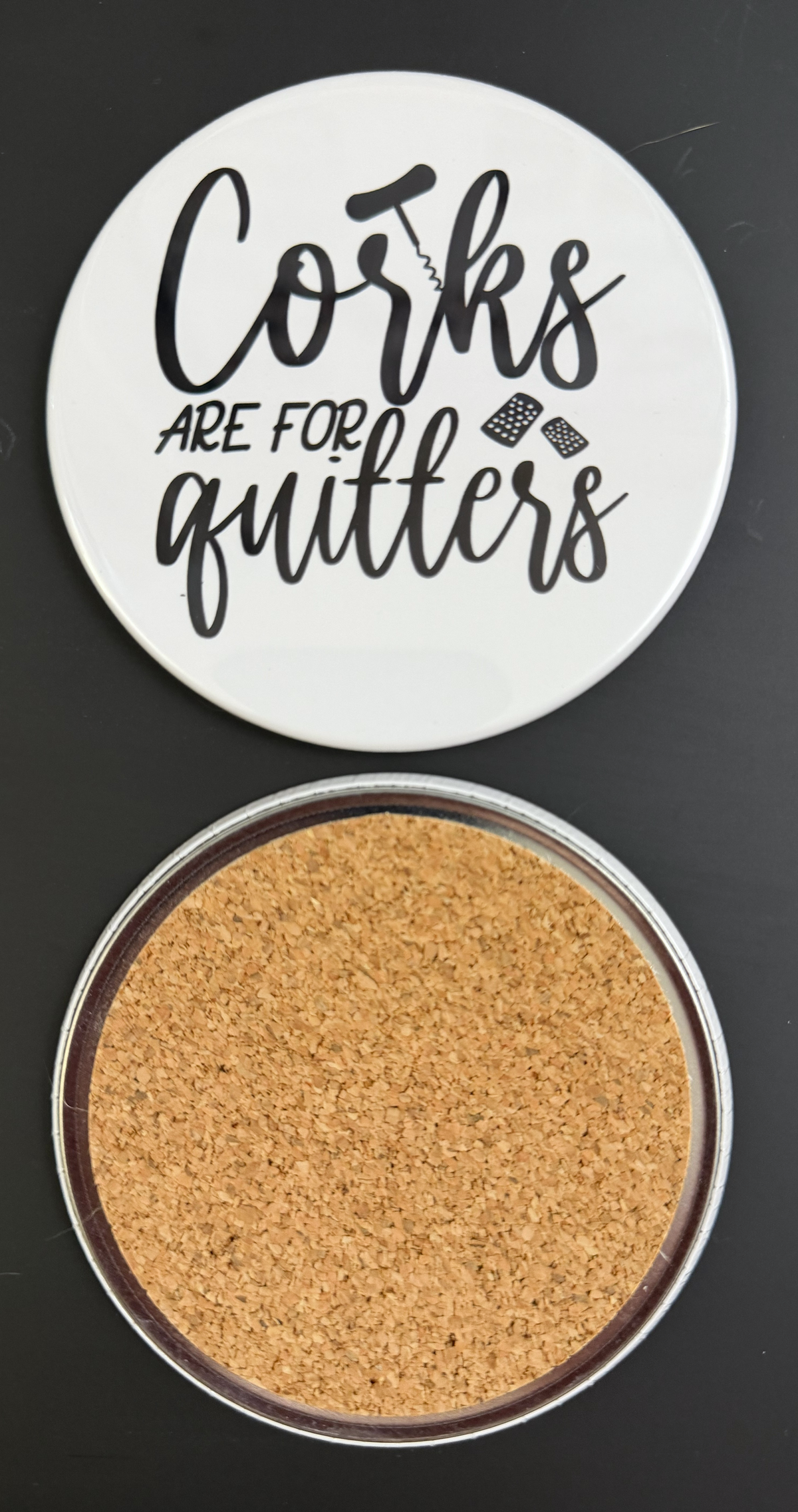 Round white drink coaster with cork backing with 'Corks are for quitters' in text black text, with black images of a corkscrew and wine corks photographed on a black background.
