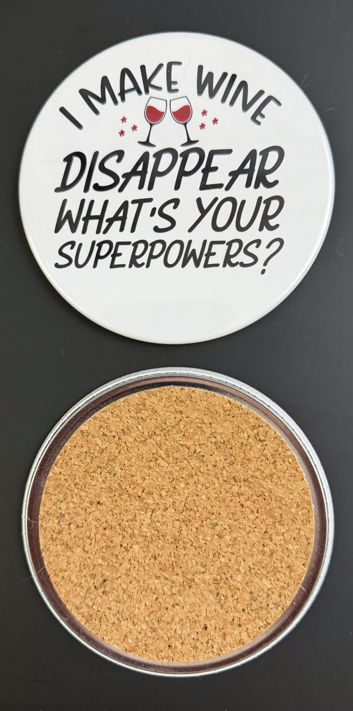 Round white drink coaster with cork backing with 'I make wine disappear, what's your superpower?' in black text and 2 glasses of red wine photographed on a black background.