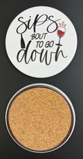 Round white drink coaster with cork backing with 'Sips 'bout to go down' in black text, with a glass of red wine and a black wine bottle, photographed on a black background.