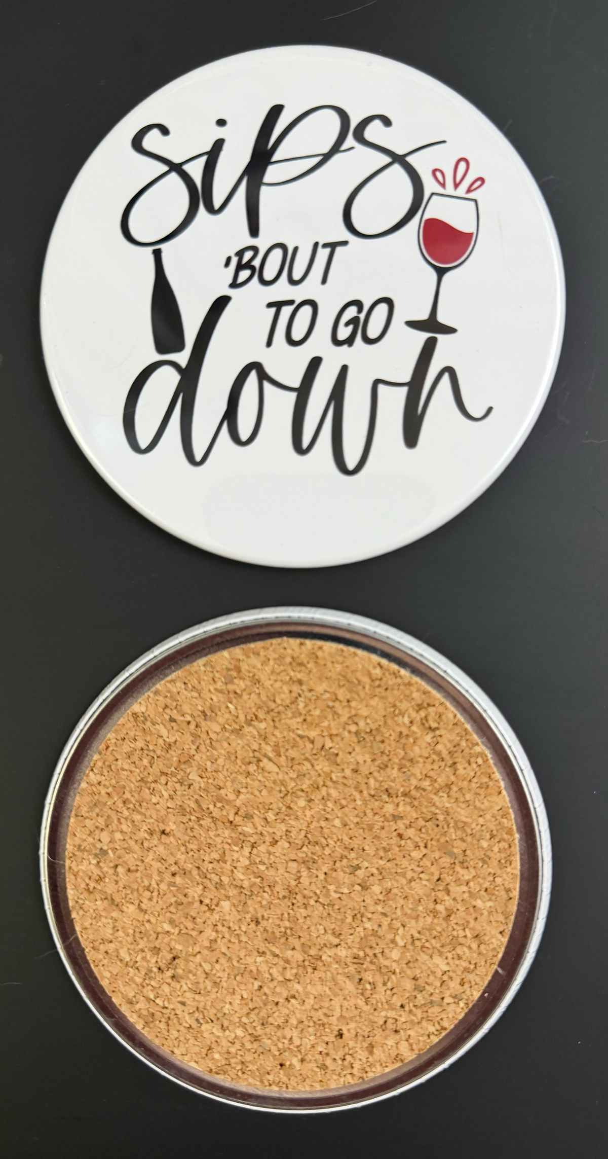 Round white drink coaster with cork backing with 'Sips 'bout to go down' in black text, with a glass of red wine and a black wine bottle, photographed on a black background.