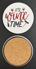 Round white drink coaster with cork backing with 'It's wine time' in red and black text, with 2 glasses of red wine, a black wine bottle and black corkscrew, photographed on a black background.