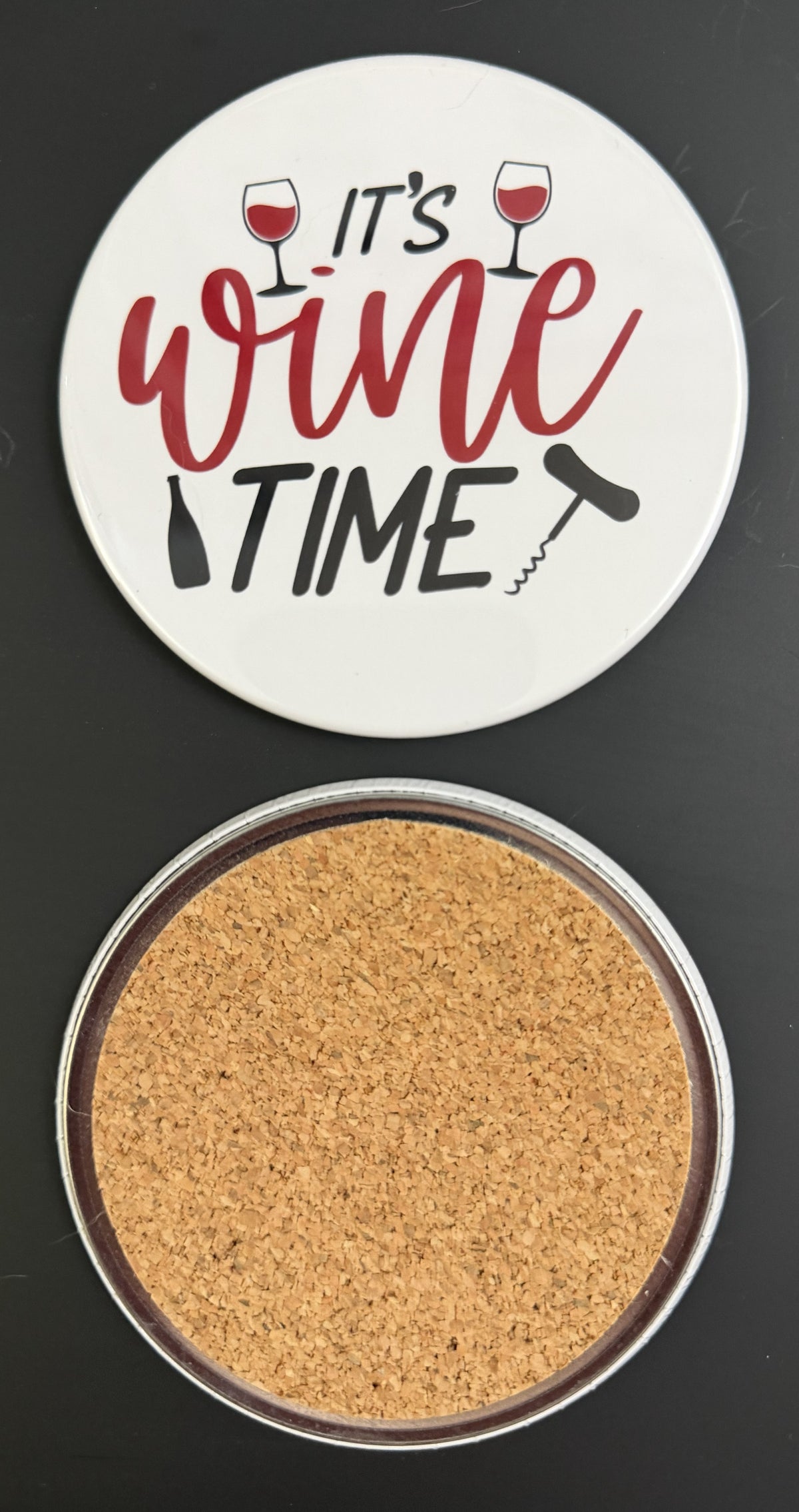 Round white drink coaster with cork backing with 'It's wine time' in red and black text, with 2 glasses of red wine, a black wine bottle and black corkscrew, photographed on a black background.