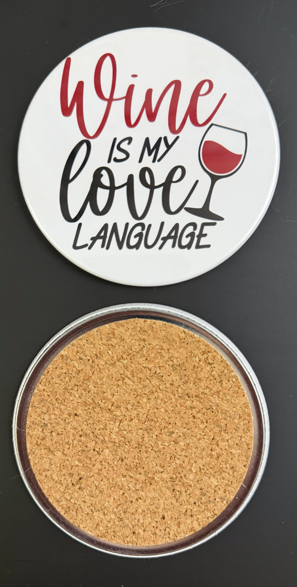 Round white drink coaster with cork backing with 'Wine is my love language' in red and black text, with a glass of red wine, photographed on a black background.