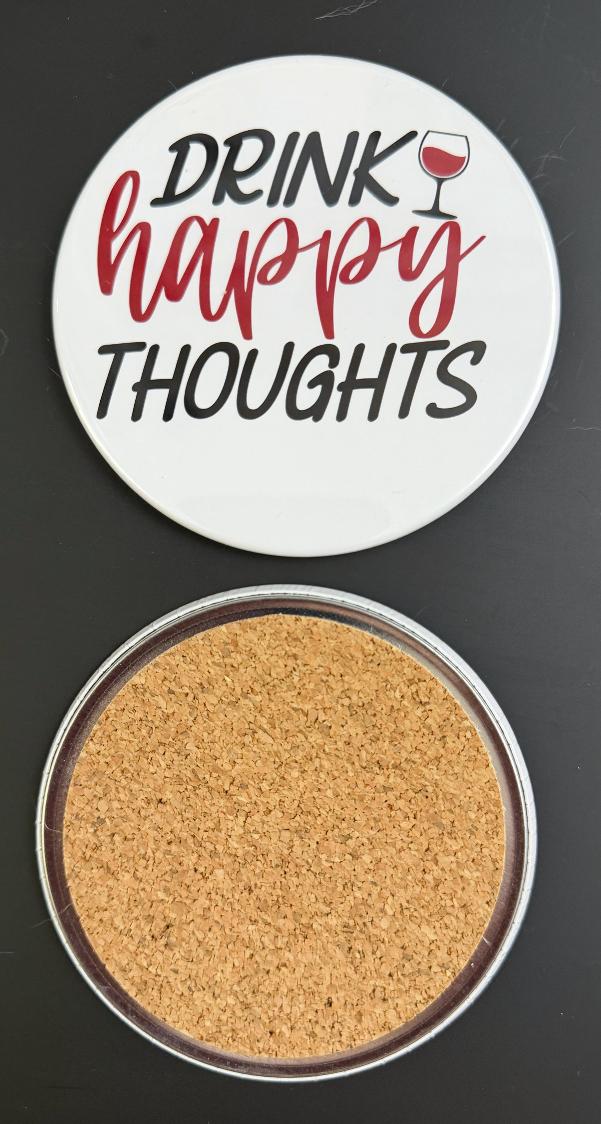 Round white drink coaster with cork backing with 'Drink Happy Thoughts' in red and black text, with a glass of red wine, photographed on a black background.