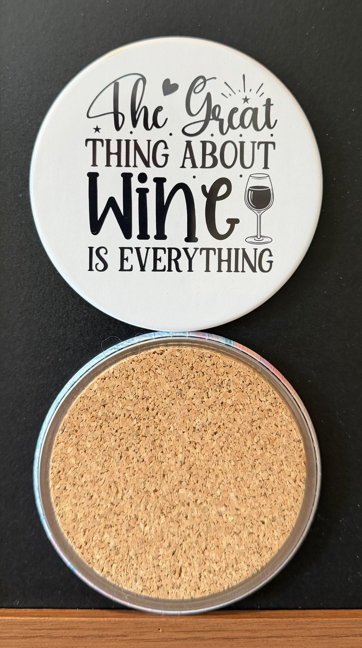 White round drink coaster with a cork backing with a black image of a black wine glass and black heart with text 'The great thing about wine is everything' on a white background.  This photo was taken on a black background.