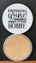 White round drink coaster with a cork backing with small black images of a wine glass and stars with text 'Drinking wine is not my habit, it's my hobby' on a white background.  This photo was taken on a black background.