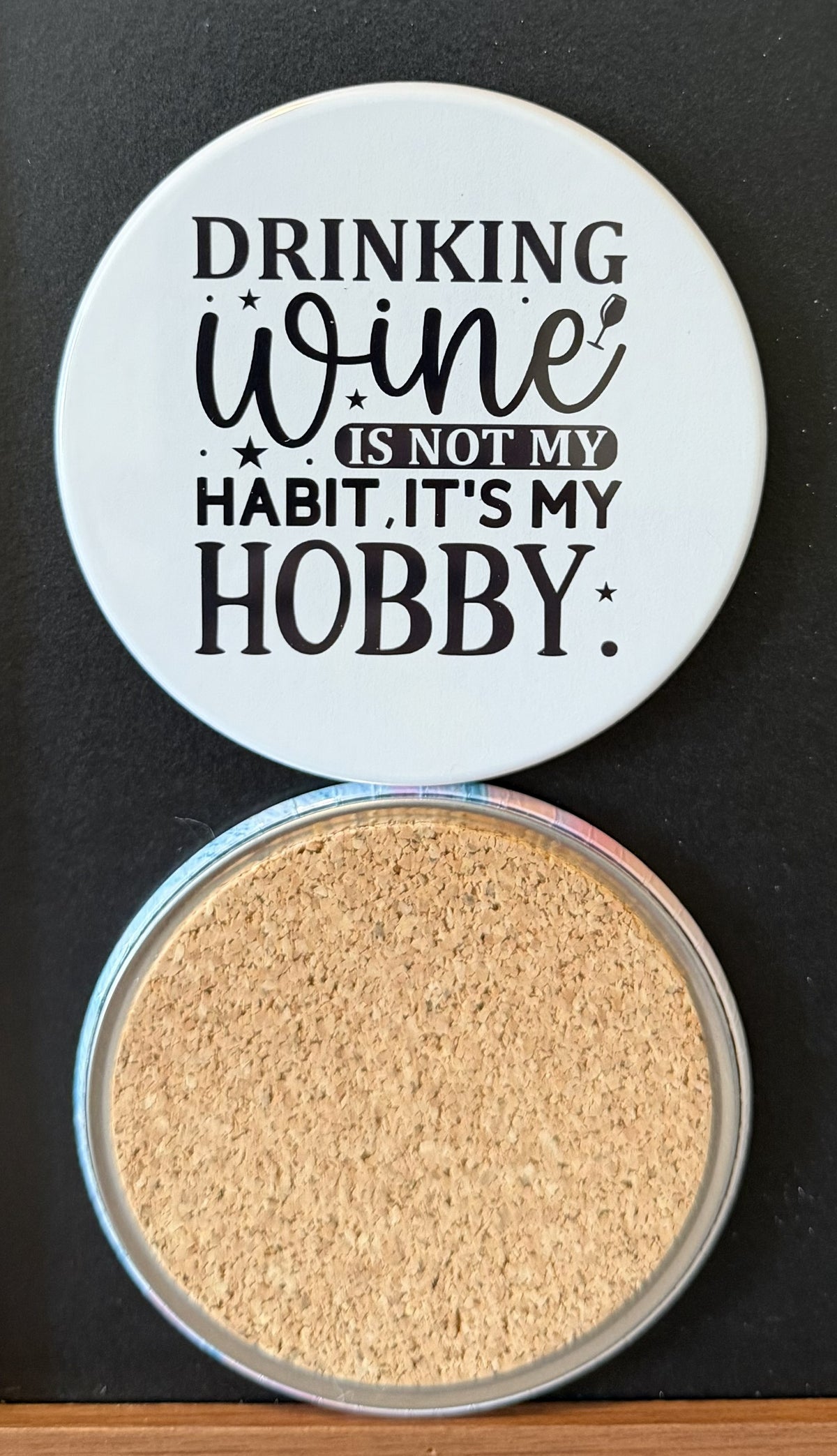 White round drink coaster with a cork backing with small black images of a wine glass and stars with text 'Drinking wine is not my habit, it's my hobby' on a white background.  This photo was taken on a black background.