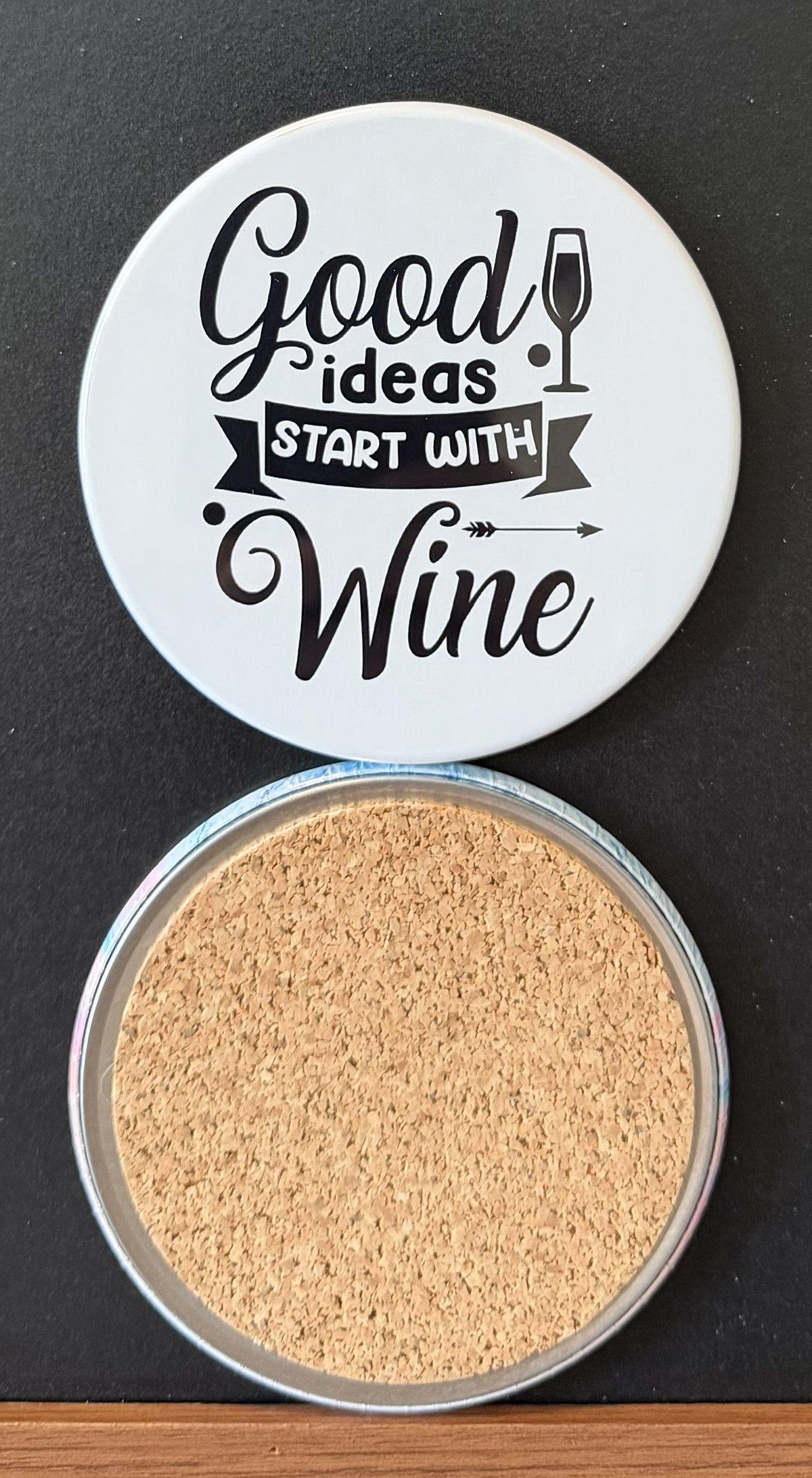 White round drink coaster with a cork backing with a black image of a WINE GLASS AND ARROW with text 'good ideas start with wine' on a white background.  This photo was taken on a black background.