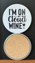 White round drink coaster with a cork backing with a black image of a WINE GLASS AND ARROW with text 'I'm on cloud wine' on a white background.  This photo was taken on a black background.