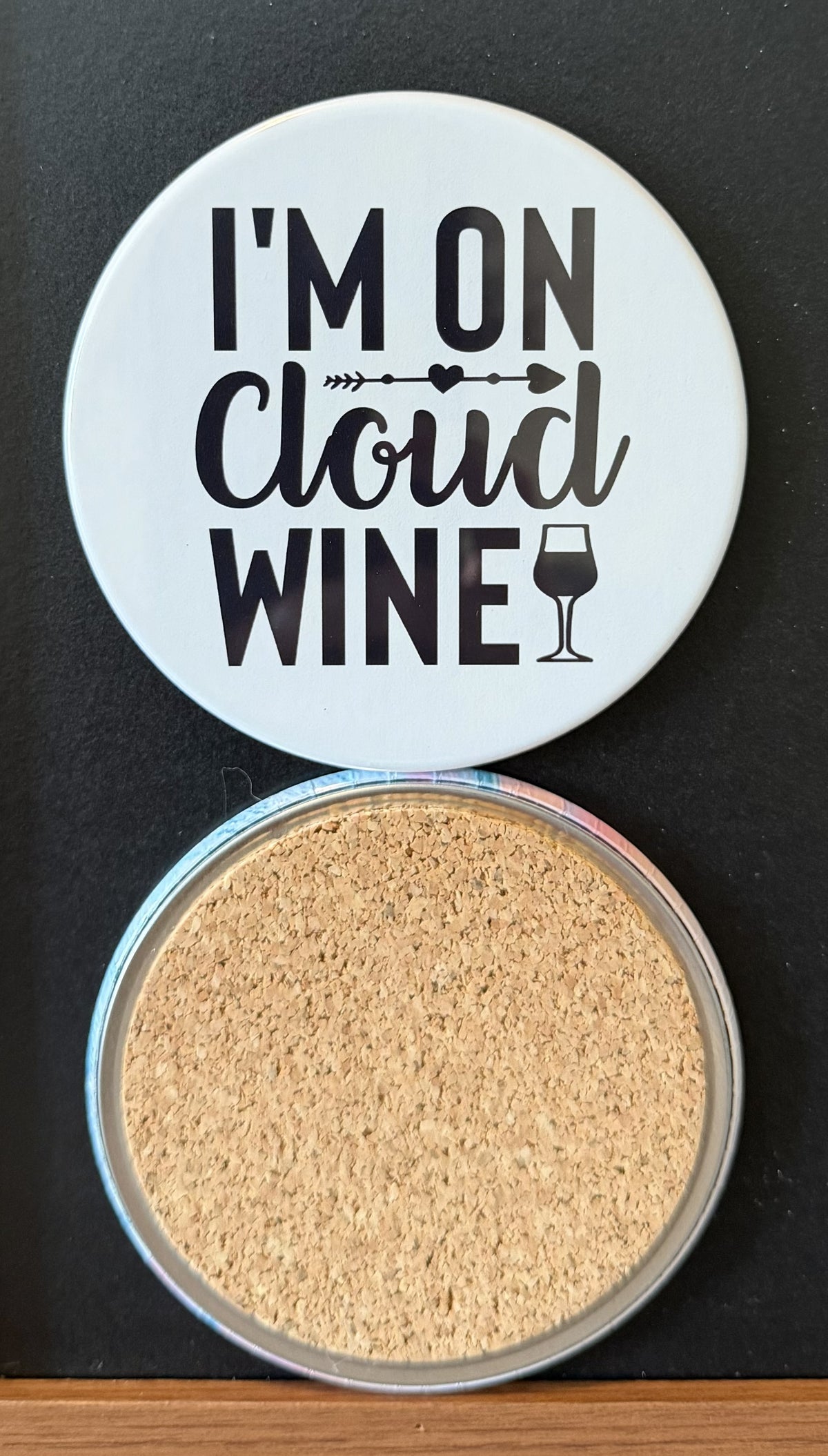 White round drink coaster with a cork backing with a black image of a WINE GLASS AND ARROW with text 'I'm on cloud wine' on a white background.  This photo was taken on a black background.