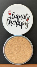White round drink coaster with a cork backing with black and red images of a wine glass and bottle with text 'liquid therapy' on a white background.  This photo was taken on a black background.