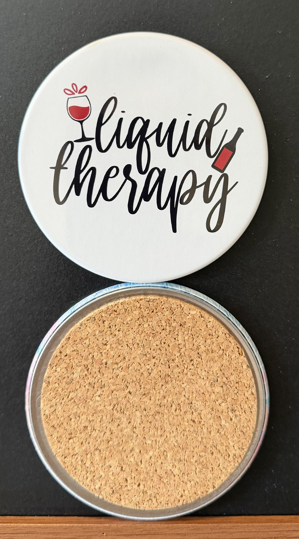 White round drink coaster with a cork backing with black and red images of a wine glass and bottle with text 'liquid therapy' on a white background.  This photo was taken on a black background.