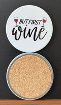White round drink coaster with a cork backing with two images of red wine glasses with text 'but first, wine' on a white background.  This photo was taken on a black background.