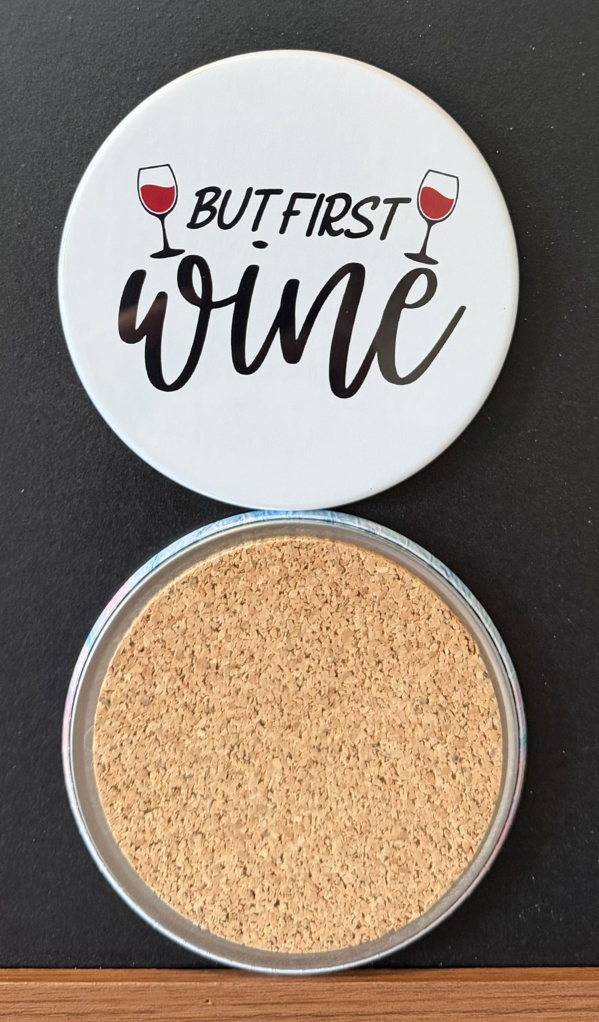 White round drink coaster with a cork backing with two images of red wine glasses with text 'but first, wine' on a white background.  This photo was taken on a black background.