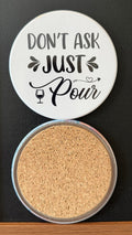 White round drink coaster with a cork backing with black images of a wine glass with wine and a decorative heart with text 'don't ask, just pour' on a white background.  This photo was taken on a black background.