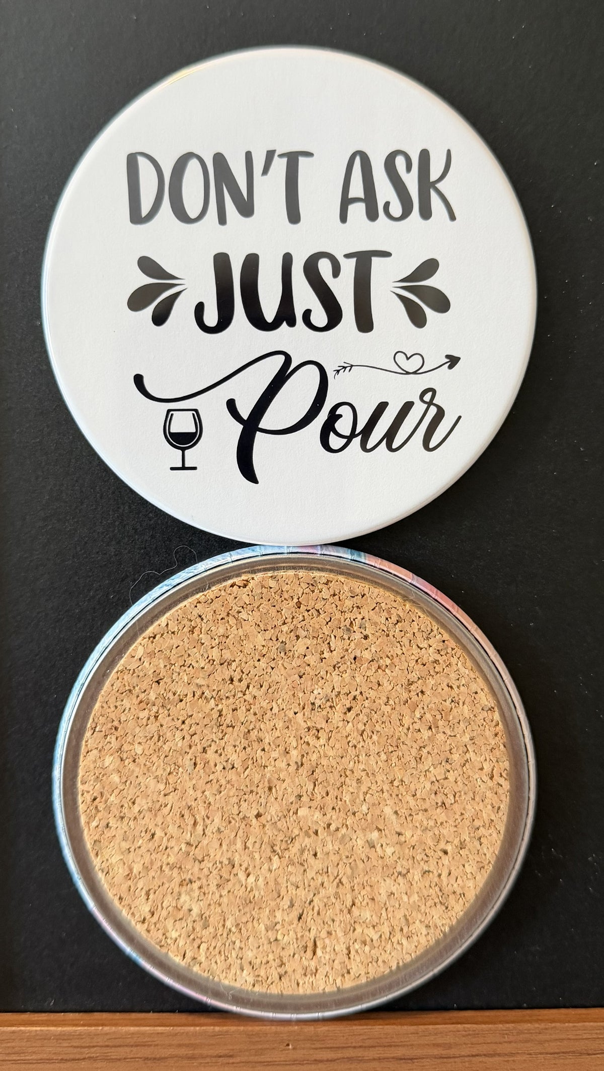 White round drink coaster with a cork backing with black images of a wine glass with wine and a decorative heart with text 'don't ask, just pour' on a white background.  This photo was taken on a black background.