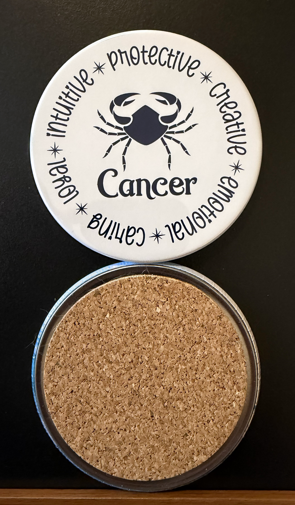 Round white drink coaster with cork backing representing the Cancer zodiac design displaying different characteristics in dark navy blue text associated with the Cancer zodiac sign, on a black background.