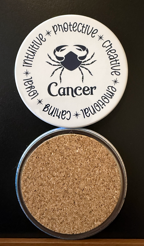 Round white drink coaster with cork backing representing the Cancer zodiac design displaying different characteristics in dark navy blue text associated with the Cancer zodiac sign, on a black background.